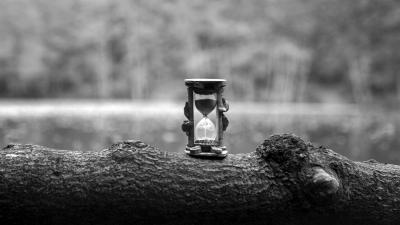 A black and white photograph of an hourglass standing on a tree log.